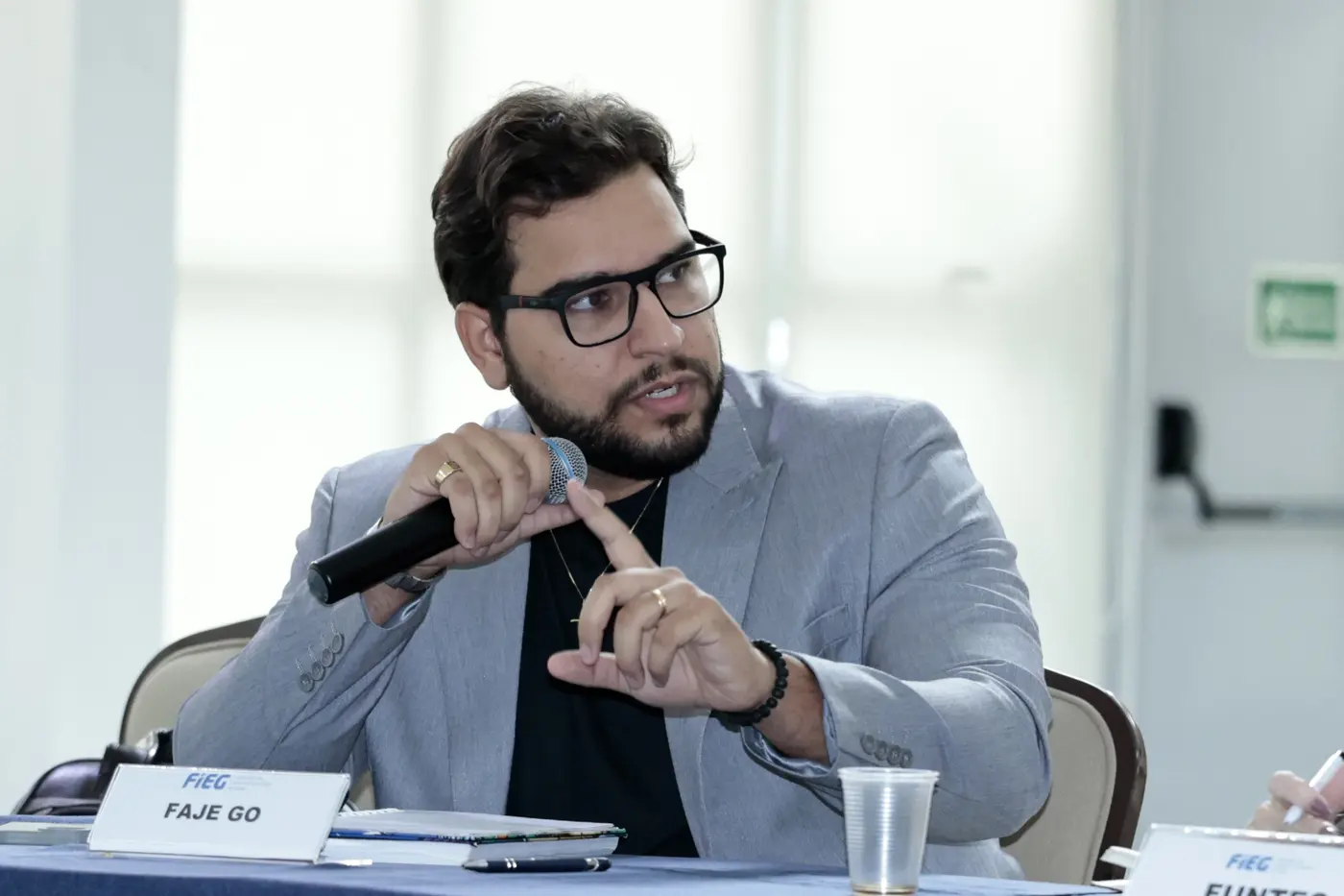 Pedro Lucas Andrade Bueno, presidente da FAJE Goiás, fala ao microfone na 23ª reunião do Conselho Gestor do Pacto Goiás pela Inovação, na Casa da Indústria, com a placa FAJE GO à sua frente na mesa.